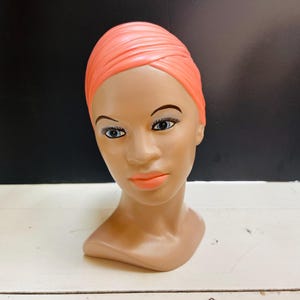 May include: A ceramic bust of a woman with a pink headscarf. The bust has a realistic face with brown eyes and a light brown complexion.