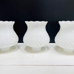 Puede incluir: Tres pantallas de lámpara de vidrio blanco con textura de clavo y bordes festoneados. Las pantallas están dispuestas en fila sobre una superficie blanca sobre un fondo negro. Tienen una estética clásica y vintage.