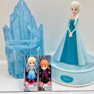 May include: A collection of Frozen II themed toys. Includes a large Elsa figurine in a blue dress, two small dolls in boxes, and a blue ice castle structure. The Elsa figurine is on a light blue base with the name "Elsa" on it.