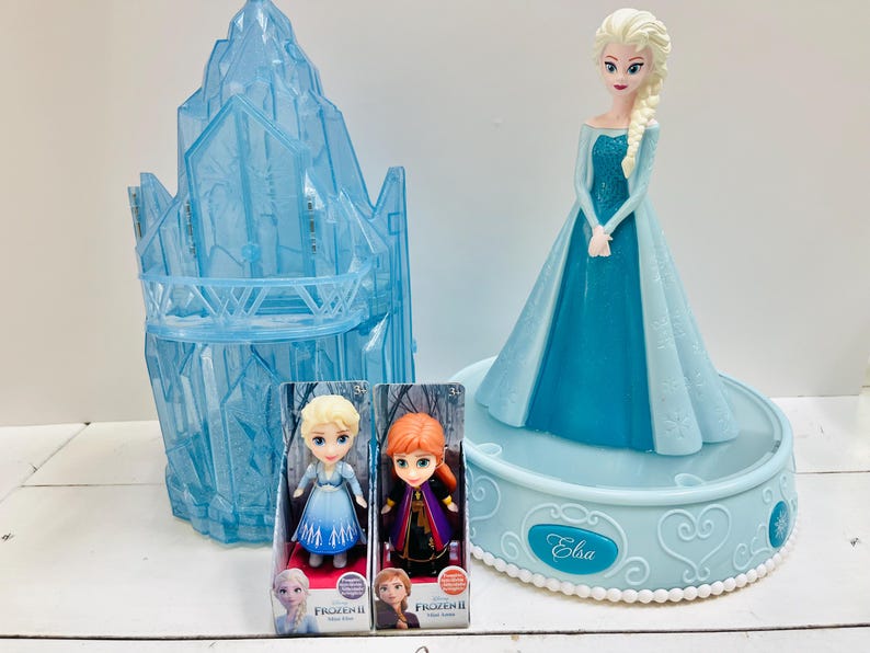 May include: A collection of Frozen II themed toys. Includes a large Elsa figurine in a blue dress, two small dolls in boxes, and a blue ice castle structure. The Elsa figurine is on a light blue base with the name "Elsa" on it.