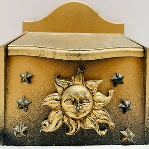 以下が含まれることがあります： 金の仕上げと顔のデザインが施された太陽が描かれた木製の箱。箱は黒い星で飾られています。
