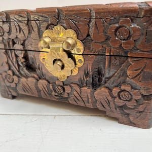 May include: A wooden box with a carved floral design and a brass latch. The box is dark brown and has a rustic appearance.