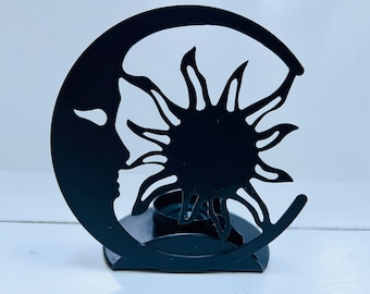 May include: Black metal candle holder shaped like a sun and moon. The sun has a stylized design with rays and the moon has a crescent shape.