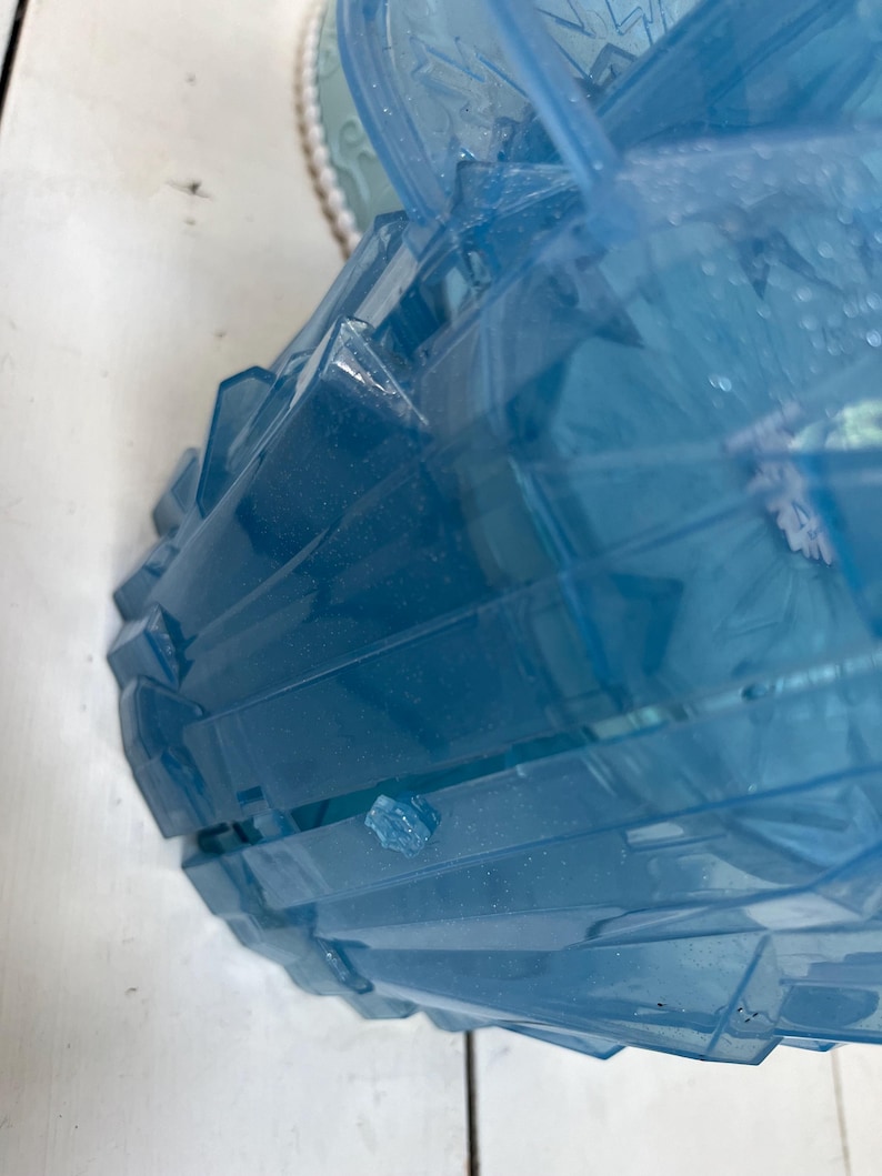 May include: A partially visible, light blue, plastic container with a geometric, faceted design. The container has a lid with a snowflake design. The container is on a white surface.