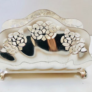 May include: Silver napkin holder with a floral design. The holder has a curved top and three floral clusters. The base has small, curved feet. The napkin holder is made of a shiny, reflective metal.