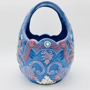 May include: A blue ceramic basket with a handle, featuring ornate embossed floral and scroll designs. The basket has a textured surface and is decorated with red, white, and yellow accents. The basket is likely vintage or antique.
