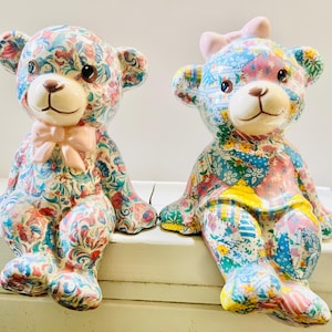 Puede incluir: Dos figuras de osos de peluche de cerámica con un diseño de patchwork floral en tonos azules, rosas, rojos y verdes. Un oso tiene una pajarita rosa, el otro un lazo rosa en la cabeza. Están sentados sobre una superficie blanca, ideales para decoración.