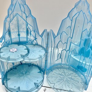 May include: A light blue plastic playset resembling an ice castle. The set features a multi-tiered structure with snowflake designs and a large, crystalline facade. The playset is designed to open and close, with a hinged door.
