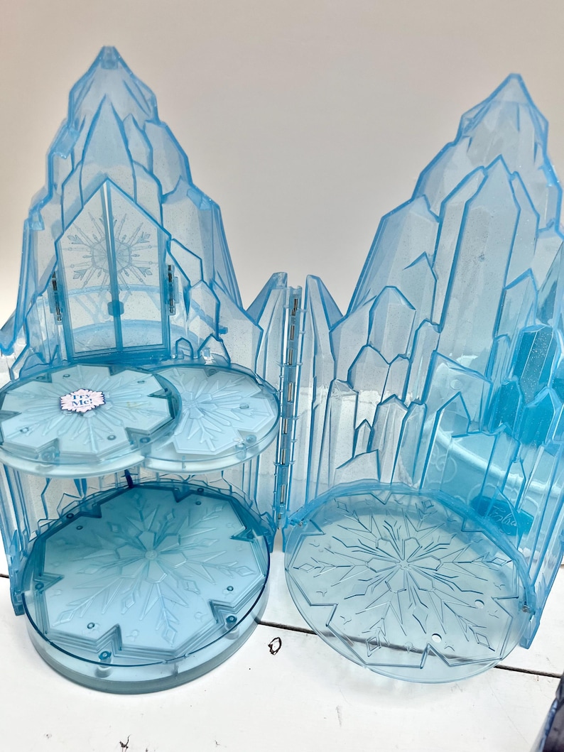 May include: A light blue plastic playset resembling an ice castle. The set features a multi-tiered structure with snowflake designs and a large, crystalline facade. The playset is designed to open and close, with a hinged door.