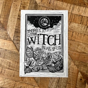 Op de afbeelding: Zwart-witte houtsnede met de tekst "There's a little witch in all of us". De print toont een halve maan, sterren, wolken, rozen en een citroen.