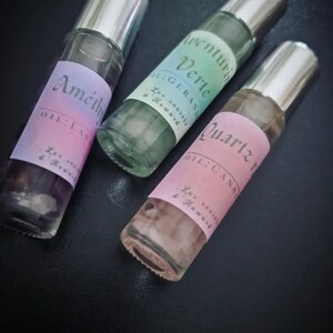 Peut inclure: Trois flacons en verre avec des bouchons argentés et des étiquettes. Les étiquettes sont rose, verte et bleue avec du texte qui dit "Amethyst Oil Lavender", "Verte Oil Geranium" et "Quartz Oil Canni", respectivement. Les flacons contiennent des cristaux et un liquide clair.