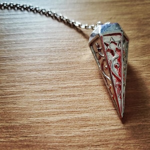 Peut inclure: Un pendule en métal argenté et rouge avec une étoile et d'autres motifs décoratifs. Le pendule est suspendu à une chaîne argentée.