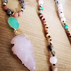 Peut inclure: Gros plan sur trois colliers de perles. Un collier présente un grand pendentif en forme de pointe de flèche rose clair avec une bordure dorée. Les autres colliers ont des perles de couleurs variées, dont rose, gris et or. Les colliers sont sur une surface en bois.
