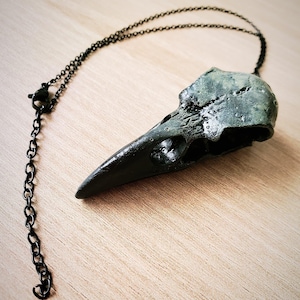 Peut inclure: Un collier pendentif en forme de crâne de corbeau. Le crâne est de couleur vert foncé avec une surface texturée, et le bec est noir. La chaîne du collier est noire et possède une fermeture à fermoir mousqueton. Le pendentif est présenté sur une surface en bois brun clair.