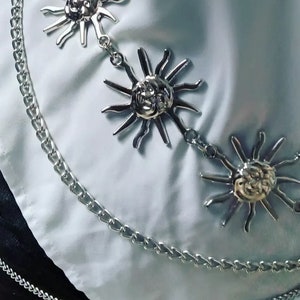 Peut inclure: Une ceinture en chaîne argentée avec plusieurs breloques en forme de soleil. La chaîne est composée de deux styles de maillons différents.