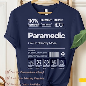 May include: Navy blue t-shirt with the word "Paramedic" in white block letters. The shirt includes the text "Life On Standby Mode" and other graphics. The shirt is hanging on a wooden hanger.