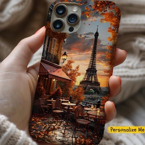 May include: A phone case with a colorful image of the Eiffel Tower in Paris, France. The image features a cafe with tables and chairs, autumn leaves, and a sunset sky. The case is designed for an iPhone with a camera lens cutout.