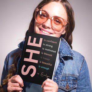 May include: A black journal with the word "SHE" in pink letters and the text "is confident, is strong, is motivated, is blessed, is happy, is beautiful, is enough, is Evelyn" in white and blue letters.