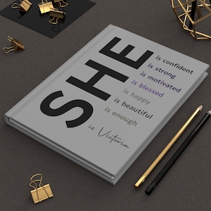 May include: A gray journal with the word "SHE" in large black letters. The text "is confident, is strong, is motivated, is blessed, is happy, is beautiful, is enough, is Victoria" is printed in black and purple on the cover.