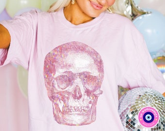 May include: Pink t-shirt with a large iridescent glitter skull graphic. The shirt is worn by a person with blonde hair and a sparkly headband. The background is a wall of balloons.