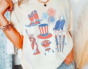 May include: A white t-shirt with a patriotic design featuring an eagle, fireworks, a grill, a cowboy boot, the Statue of Liberty, and a top hat. The text "IVORY" is in a heart shape in the top right corner of the image.