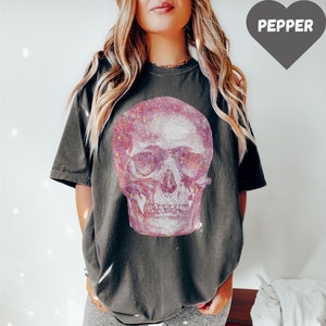 Op de afbeelding: Een oversized donkergrijs T-shirt met een roze glitter schedel print. Het shirt wordt gedragen door een persoon met lang blond haar. De tekst "PEPPER" is zichtbaar in een hartvormige afbeelding.