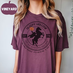 May include: A purple t-shirt with a black graphic of a headless horseman riding a horse. The graphic includes the text "Sleepy Hollow", "Est 1790", "Dead and Breakfast", and "Make Sure to Book a Head".