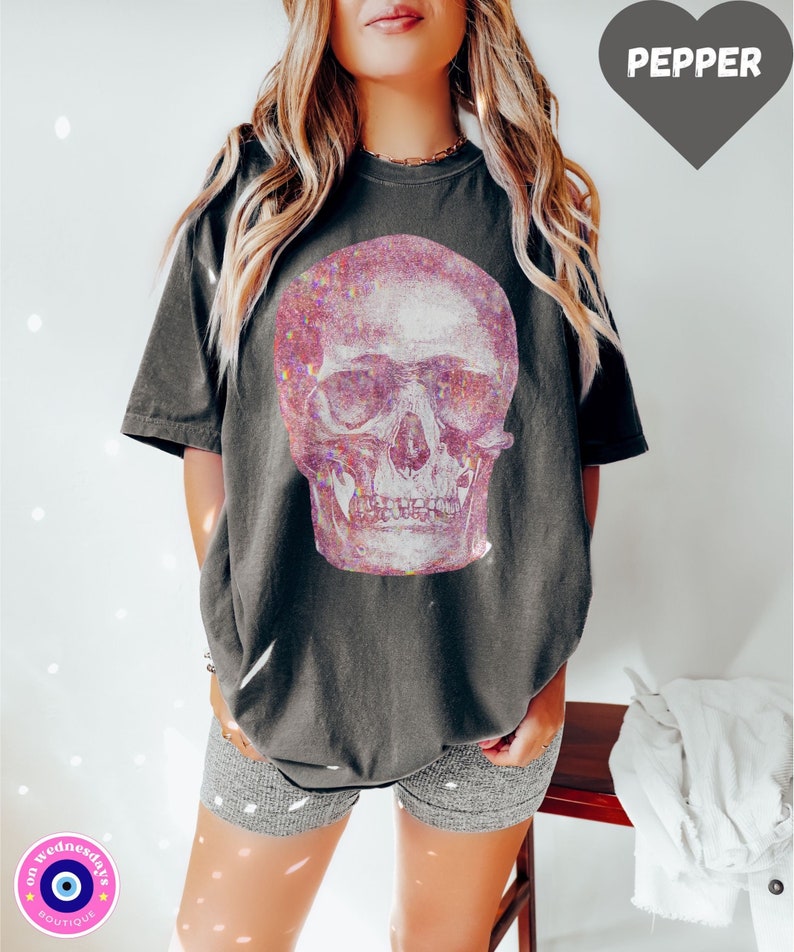 Op de afbeelding: Een oversized donkergrijs T-shirt met een roze glitter schedel print. Het shirt wordt gedragen door een persoon met lang blond haar. De tekst "PEPPER" is zichtbaar in een hartvormige afbeelding.