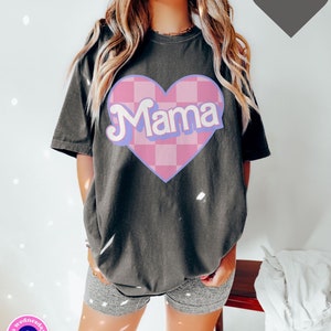Puede incluir: Una camiseta gris oscuro con un coraz&oacute;n a cuadros rosa y blanco que dice "Mama" en letras de color morado claro. La camiseta es de gran tama&ntilde;o y tiene un corte holgado.