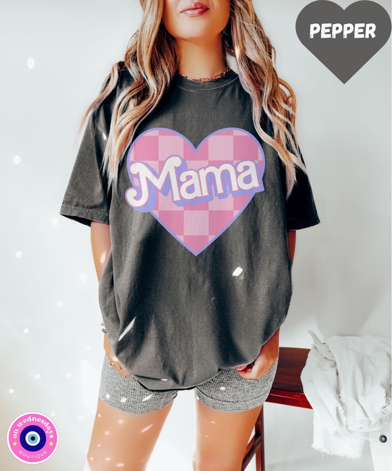 Puede incluir: Una camiseta gris oscuro con un coraz&oacute;n a cuadros rosa y blanco que dice "Mama" en letras de color morado claro. La camiseta es de gran tama&ntilde;o y tiene un corte holgado.