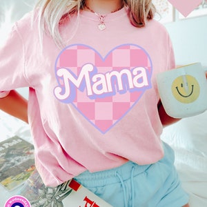 Puede incluir: Camiseta rosa con un coraz&oacute;n a cuadros y la palabra "Mama" en una fuente retro.