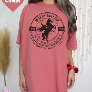 May include: A pink t-shirt with a black and white graphic of a headless horseman riding a horse. The graphic is in a circle with the text "Sleepy Hollow" at the top, "Est 1790" on the right, "Dead and Breakfast" at the bottom left, and "Make Sure to Book a Head" at the bottom right.