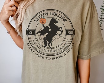 May include: Khaki t-shirt with a black and white graphic of a headless horseman riding a horse with a pumpkin in its hand. The graphic is in a circle with the text "Sleepy Hollow" at the top, "Est 1790" on the right, "Dead and Breakfast" at the bottom left, and "Make Sure to Book a Head" at the bottom right.