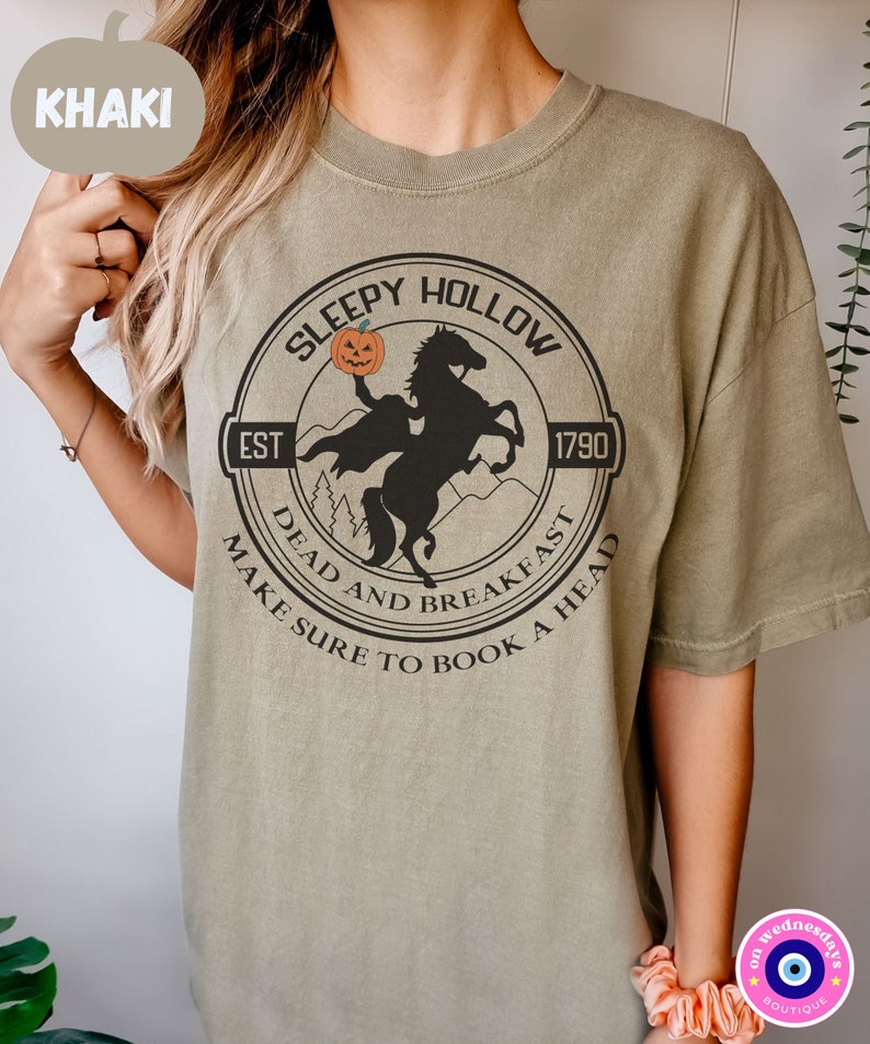 May include: Khaki t-shirt with a black and white graphic of a headless horseman riding a horse with a pumpkin in its hand. The graphic is in a circle with the text "Sleepy Hollow" at the top, "Est 1790" on the right, "Dead and Breakfast" at the bottom left, and "Make Sure to Book a Head" at the bottom right.