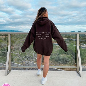 K&ouml;nnte beinhalten: Ein dunkelbrauner Kapuzenpullover mit dem Text "THE TIMING OF YOUR LIFE WILL MAKE SENSE ONE DAY" auf dem R&uuml;cken. Der Kapuzenpullover wird von einer Person getragen, die auf einem Balkon mit Blick auf eine Landschaft steht. Wei&szlig;e Turnschuhe vervollst&auml;ndigen das Outfit.