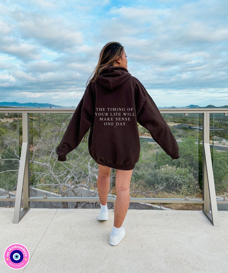 K&ouml;nnte beinhalten: Ein dunkelbrauner Kapuzenpullover mit dem Text "THE TIMING OF YOUR LIFE WILL MAKE SENSE ONE DAY" auf dem R&uuml;cken. Der Kapuzenpullover wird von einer Person getragen, die auf einem Balkon mit Blick auf eine Landschaft steht. Wei&szlig;e Turnschuhe vervollst&auml;ndigen das Outfit.