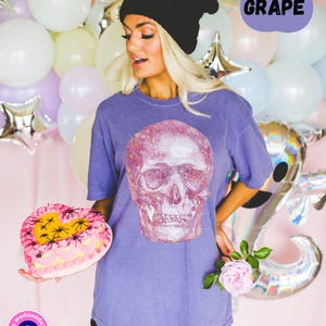 Op de afbeelding: Een vrouw draagt een paars T-shirt met een glitter schedel print, een zwarte beanie en een zwarte short. Ze houdt een roze hartvormige taart vast met zwarte spinnen erop. De tekst "GRAPE" staat in een spraakballon in de rechterbovenhoek van de afbeelding.