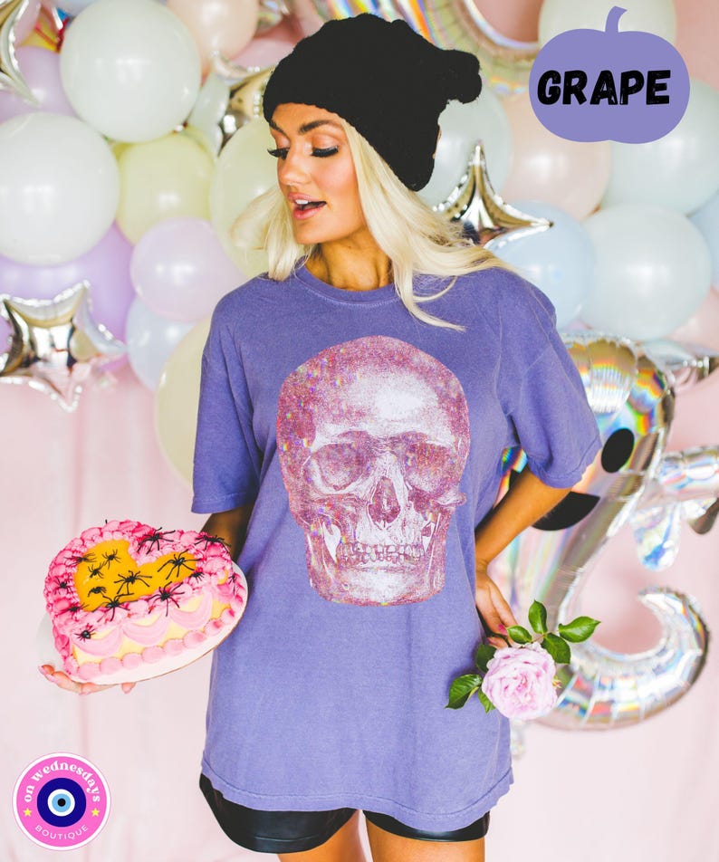Op de afbeelding: Een vrouw draagt een paars T-shirt met een glitter schedel print, een zwarte beanie en een zwarte short. Ze houdt een roze hartvormige taart vast met zwarte spinnen erop. De tekst "GRAPE" staat in een spraakballon in de rechterbovenhoek van de afbeelding.