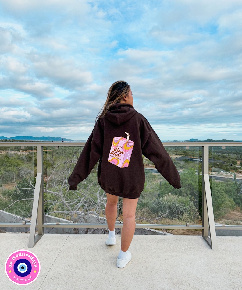 May include: A brown hoodie with a pink and yellow graphic of a juice box on the back. The juice box has the text "Boys Toys" on it. The hoodie is being worn by a person standing on a balcony overlooking a view of mountains and a cloudy sky.