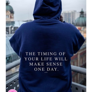 K&ouml;nnte beinhalten: Ein marineblauer Hoodie mit dem wei&szlig;en Text "THE TIMING OF YOUR LIFE WILL MAKE SENSE ONE DAY." Die Kapuzenjacke wird von einer Person getragen, die dem Betrachter den R&uuml;cken zukehrt, mit einer verschwommenen Stadtansicht im Hintergrund.