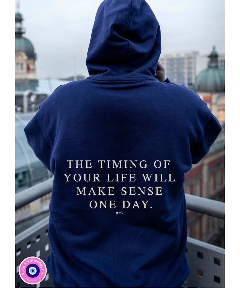 K&ouml;nnte beinhalten: Ein marineblauer Hoodie mit dem wei&szlig;en Text "THE TIMING OF YOUR LIFE WILL MAKE SENSE ONE DAY." Die Kapuzenjacke wird von einer Person getragen, die dem Betrachter den R&uuml;cken zukehrt, mit einer verschwommenen Stadtansicht im Hintergrund.