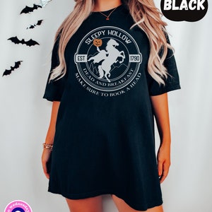 May include: Black t-shirt with a white graphic of a headless horseman riding a horse with a pumpkin in its hand. The graphic is in a circle with the text "Sleepy Hollow" and "Est 1790" above the graphic and "Dead and Breakfast Make Sure to Book a Head" below the graphic.