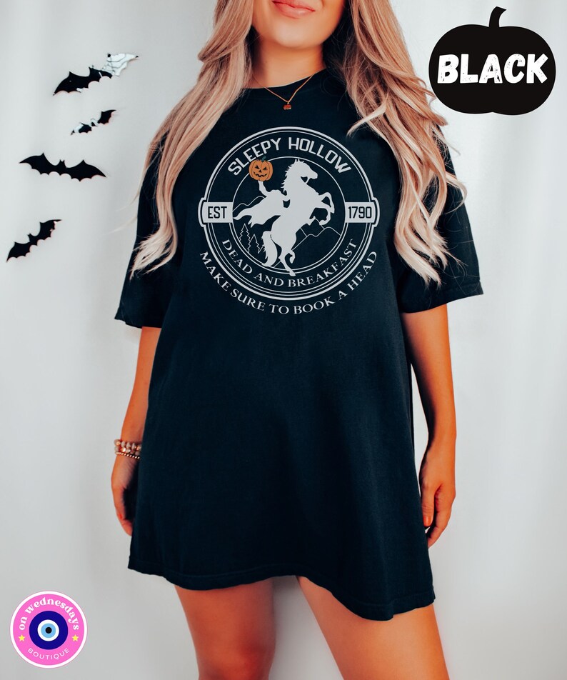 May include: Black t-shirt with a white graphic of a headless horseman riding a horse with a pumpkin in its hand. The graphic is in a circle with the text "Sleepy Hollow" and "Est 1790" above the graphic and "Dead and Breakfast Make Sure to Book a Head" below the graphic.