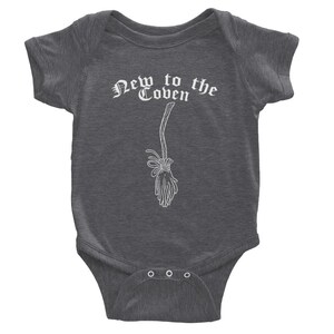 May include: Dark gray baby bodysuit with the text "New to the Coven" in a gothic font. A white illustration of a broom is below the text. The bodysuit has short sleeves and three snaps at the bottom.