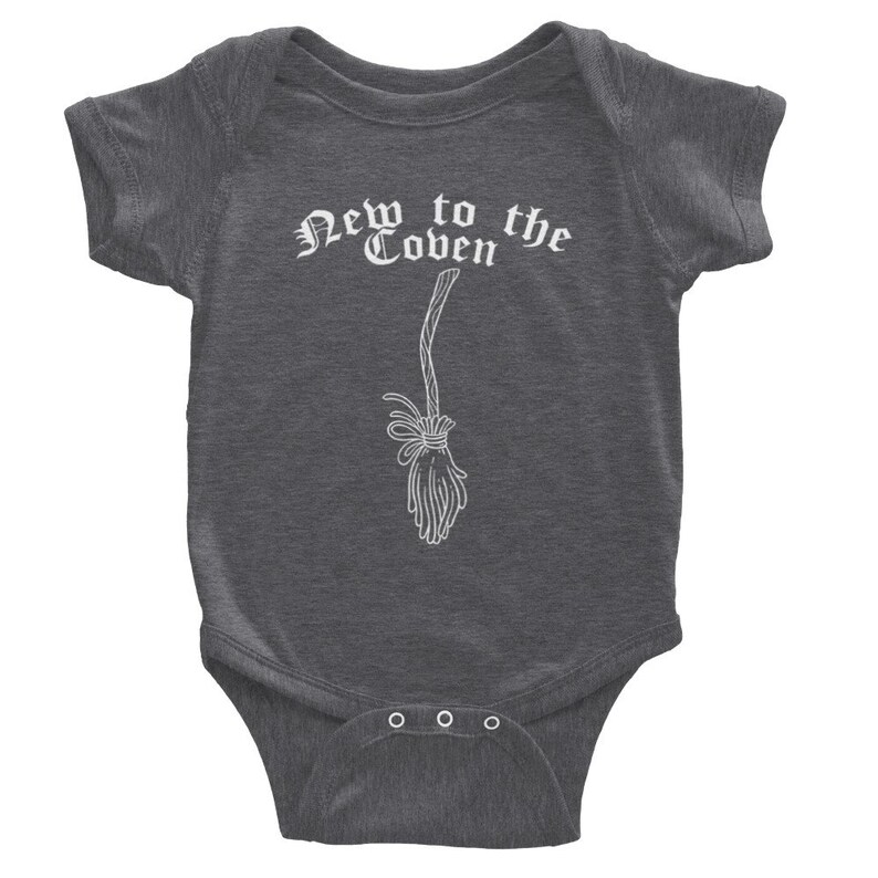 May include: Dark gray baby bodysuit with the text "New to the Coven" in a gothic font. A white illustration of a broom is below the text. The bodysuit has short sleeves and three snaps at the bottom.