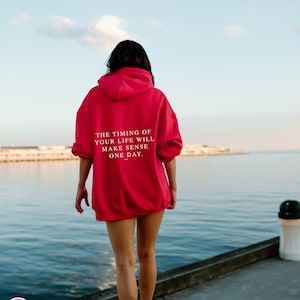K&ouml;nnte beinhalten: Ein leuchtend roter Kapuzenpullover mit dem Text "THE TIMING OF YOUR LIFE WILL MAKE SENSE ONE DAY" auf dem R&uuml;cken. Der Hoodie wird von einer Person am Wasser getragen.