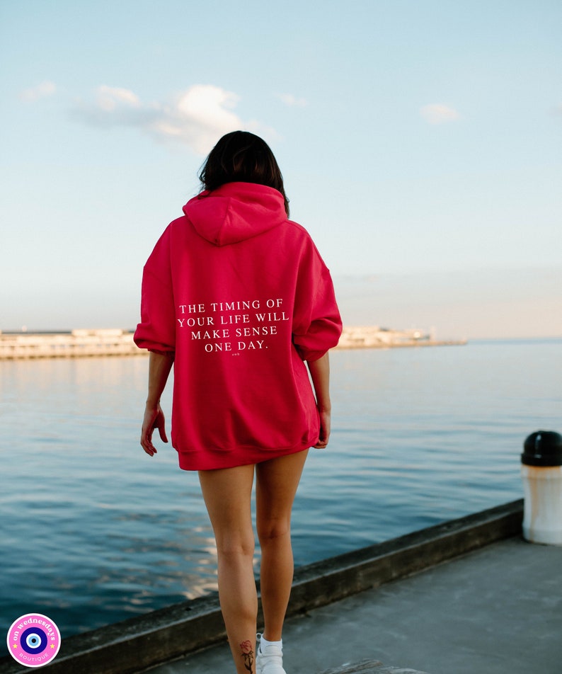 K&ouml;nnte beinhalten: Ein leuchtend roter Kapuzenpullover mit dem Text "THE TIMING OF YOUR LIFE WILL MAKE SENSE ONE DAY" auf dem R&uuml;cken. Der Hoodie wird von einer Person am Wasser getragen.