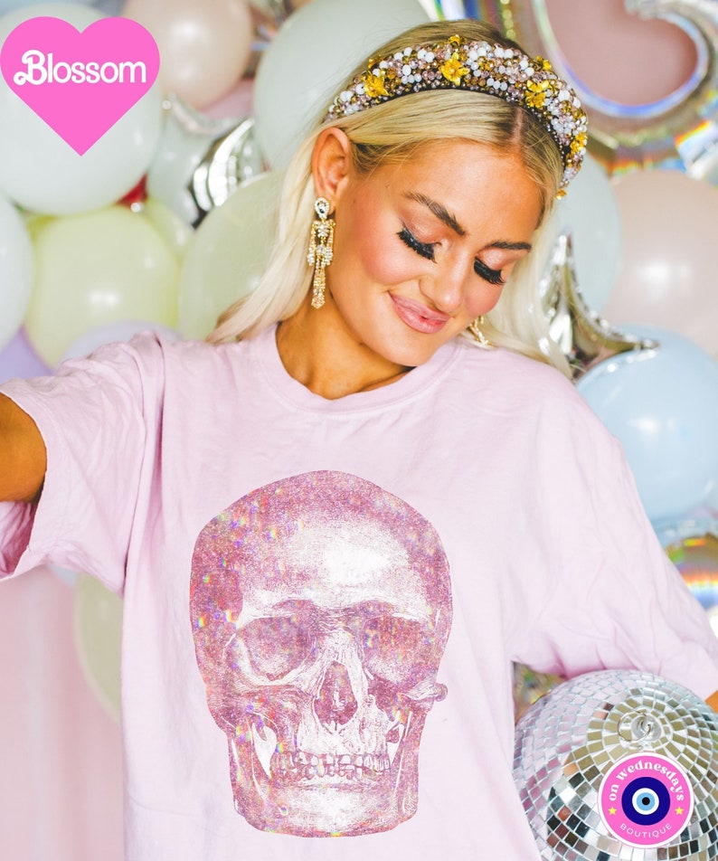 Op de afbeelding: Roze T-shirt met een grote, iriserende glitter schedel print. Het shirt wordt gedragen door een persoon met blond haar en een glinsterend hoofdband. De achtergrond is een muur van ballonnen.