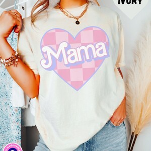 Puede incluir: Una camiseta blanca con un coraz&oacute;n a cuadros rosa y azul que dice "Mama" en una fuente retro. La camiseta es de color marfil.
