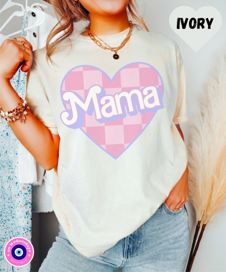 Puede incluir: Una camiseta blanca con un coraz&oacute;n a cuadros rosa y azul que dice "Mama" en una fuente retro. La camiseta es de color marfil.